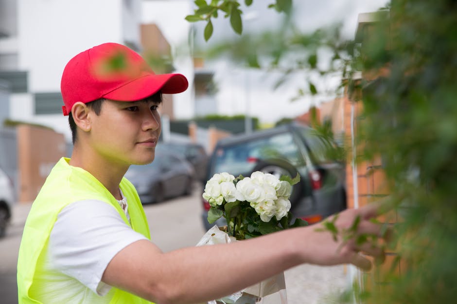 A young male florist wearing a red cap and a high-visibility yellow vest arranges a bouquet of white flowers, possibly garden roses or peonies, outdoors beside a brick wall with green foliage in the f