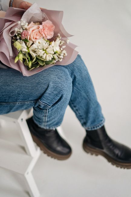 A person seated on a white stool, holding a beautifully arranged bouquet of fresh flowers wrapped in mauve and beige tissue paper. The bouquet features soft pastel pink roses, white lilies, and small
