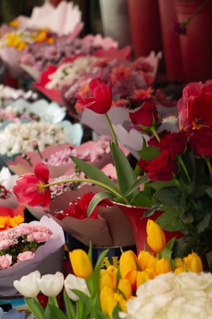 A vibrant display of fresh flowers featuring a variety of colorful blossoms arranged in bouquets at a floral shop. Prominently in the foreground, pink tulips and red and yellow freesias are placed in