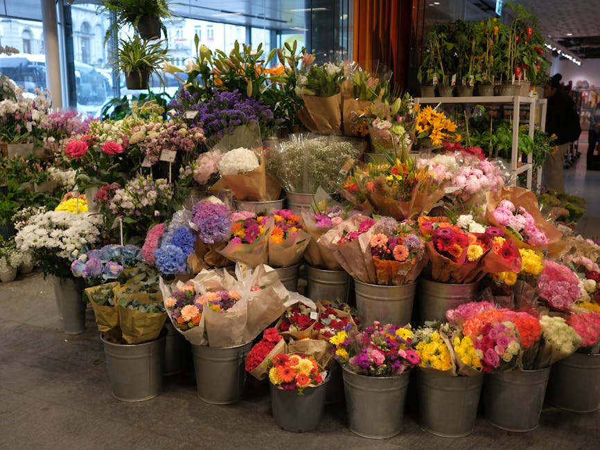 A large display of fresh flowering plants and bouquets arranged in metal buckets and paper wraps at an indoor flower shop. The arrangement includes a variety of colorful flowers such as pink gerberas,
