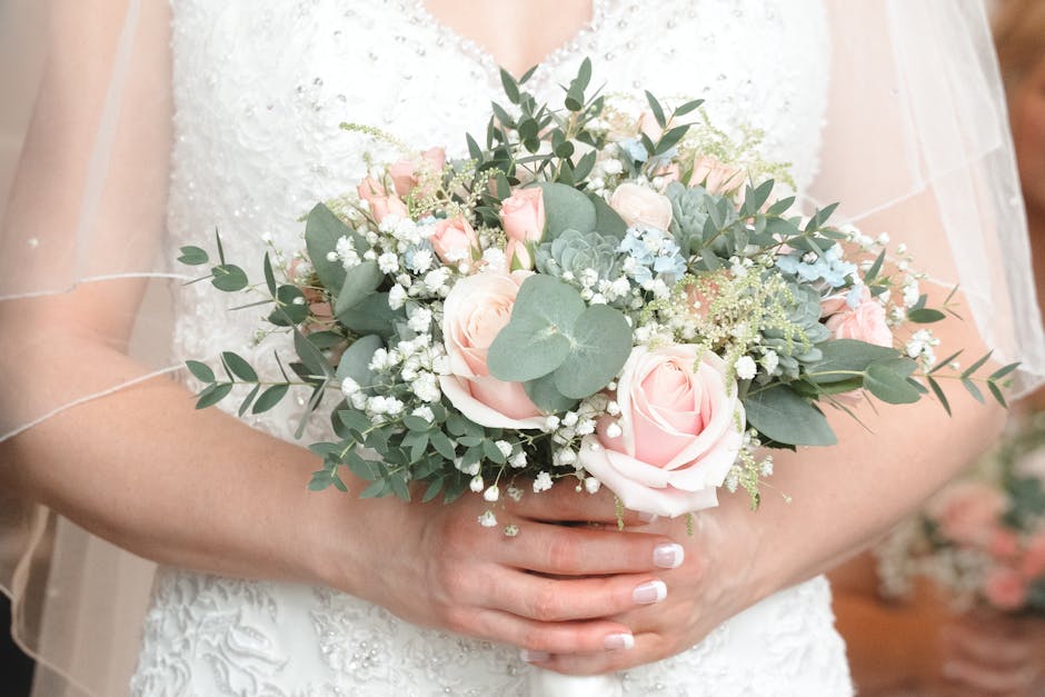 A bride in a white lace wedding dress holds a floral bouquet featuring soft pink roses, white baby's breath, silvery green eucalyptus leaves, and blue-green succulents, arranged in a rounded, elegant