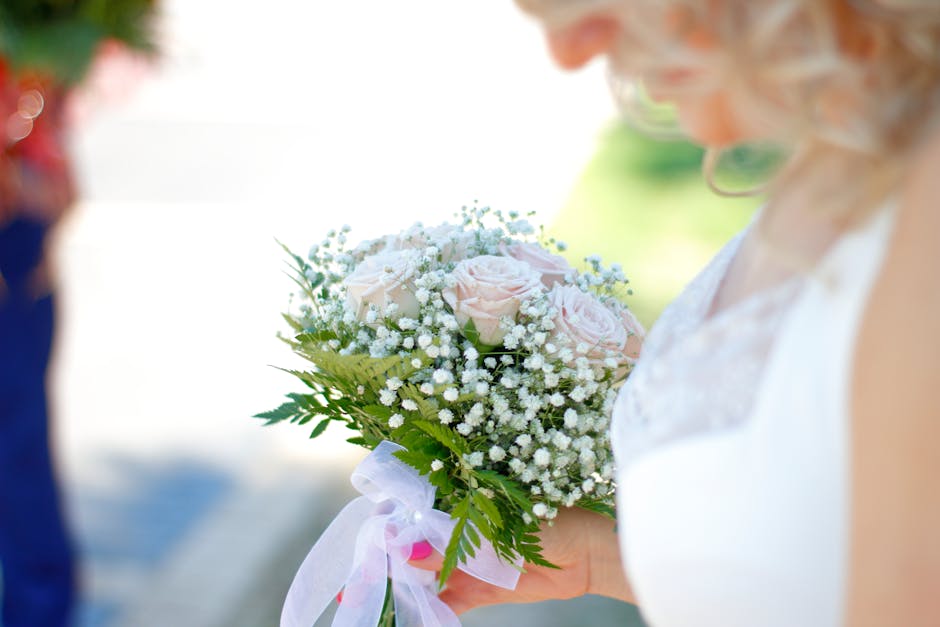A close-up of a bride holding a floral bouquet arrangement featuring pale pink roses, white baby's breath, and lush green foliage, wrapped with a light pink ribbon. The bouquet appears fresh with deli