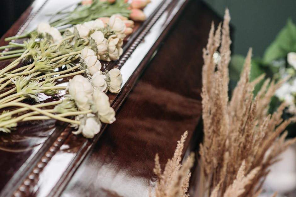 Close-up of a floral arrangement on a polished wooden surface, featuring a line of white roses with delicate petals and green stems, alongside tall, beige dried grasses with feathery plumes. The flowe