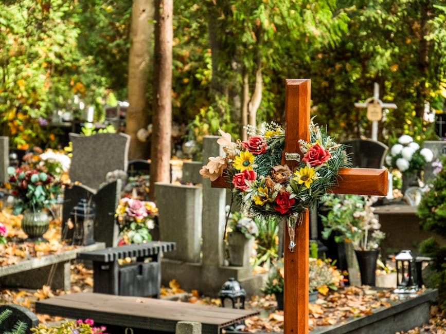 A wooden funeral cross decorated with a floral arrangement featuring red roses, yellow daisies, and pale pink flowers, surrounded by green foliage, set in a cemetery with gravestones and memorial plaq